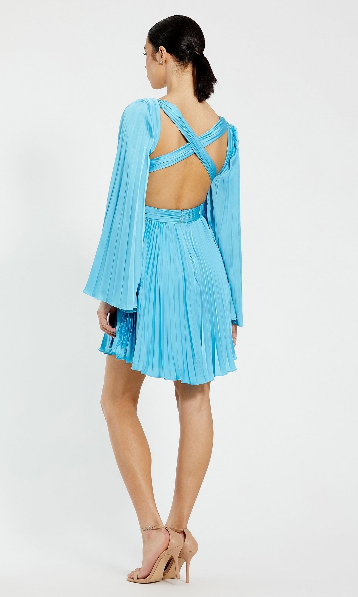 A woman faces away, wearing the Mac Duggal Long Sleeve Pleated Short Party Dress 27379 with an open back and crisscross straps, paired with nude heeled sandals.